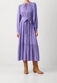 Lilla blomstret maxikjole med lange ærmer, rynket overdel, bindebånd i taljen og lagdelt nederdel. Båret med brune cowboystøvler.