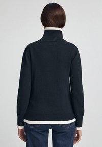 Granatowy dzianinowy sweter z wysokim kołnierzem, ściągaczami na mankietach i kremowym obrzeżem. Na lewym rękawie znajduje się naszywka.