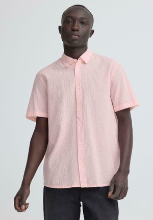 Giovane uomo con indosso una camicia a maniche corte rosa chiaro e pantaloni scuri, in piedi davanti a uno sfondo chiaro e uniforme.