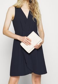 Navy jurk met een diepe V-hals en wikkeldesign, gecombineerd met een licht crèmekleurige geweven clutch, vastgehouden voor een model.