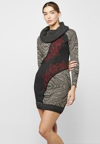 Schwarzes Kleid mit roten und beigen gemusterten Akzenten, mit einem großen Schalkragen, langen Ärmeln und einer figurbetonten Silhouette.