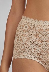 Beige spetsbrief med blommönster och geometriska mönster. Scallopade kanter vid midjan och benöppningarna förstärker dess feminina design.