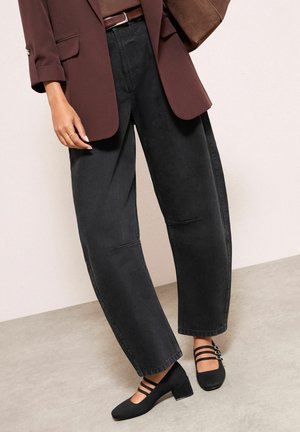 Sieviete, kas valkā melnas plaša piegriezuma džinsas, brūnu žaketi un melnas Mary Jane stila kurpes ar zemiem bloka papēžiem un dubultām siksniņām.