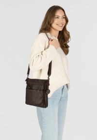 Borsa a tracolla in pelle marrone con più scomparti con zip e un charm decorativo a forma di ancora. Indossata con un maglione leggero e jeans.