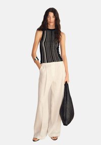 Top senza maniche a righe bianco e nero abbinato a pantaloni larghi in lino bianco. Il modello tiene una borsa intrecciata nera e indossa accessori minimalisti.