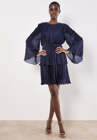 Granatowa, plisowana sukienka z długimi dzwonowymi rękawami, paskiem w talii i warstwowymi falbanami, w połączeniu z cielistymi szpilkami na kostkę.