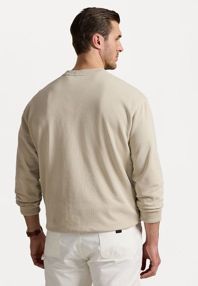 Beige pamut pulóver hosszú ujjakkal, kerek nyakkivágással és bordázott mandzsettákkal, fehér nadrággal párosítva. Minimális dizájn, látható logó nélkül.