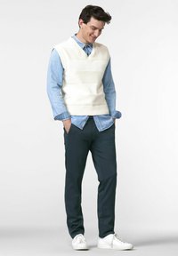 Witte gebreide vest over een lichtblauw overhemd met knopen, gecombineerd met donkergroene broek en witte sneakers. Casual outfit met gestructureerde lagen.