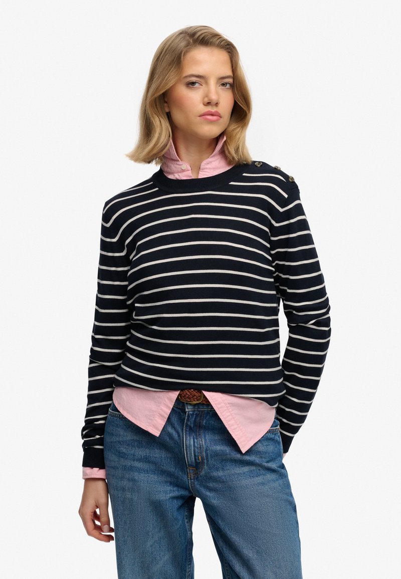 Pull rayé marine à manches longues, col rond, avec des boutons sur les épaules, superposé à une chemise à col rose clair, associé à un jean bleu.