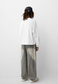 White oversized sweatshirt, loose-fitting faded gray denim trousers, and white sneakers. Dark cap with text on the back.