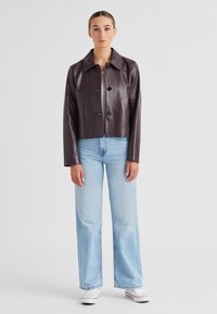 Gekürzte, dunkelviolette Lederjacke mit Knopfverschluss, spitzem Kragen und langen Ärmeln. Kombiniert mit hellblauen, weit geschnittenen Jeans und weißen Sneakers.