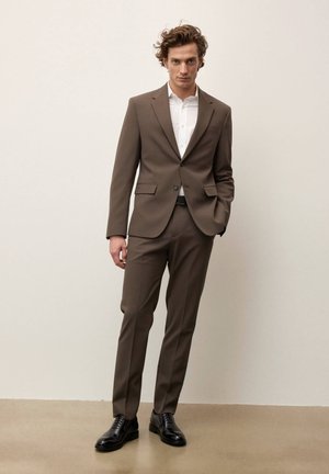 Mand med krøllet hår iført brun jakkesæt, hvid skjorte, sort bælte og sorte dresssko, stående med den ene hånd i lommen.