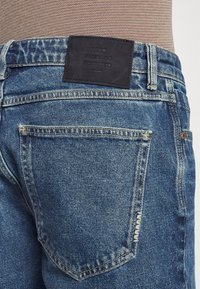 Jeans de mezclilla azul con un acabado texturizado, que cuentan con un parche de marca de cuero negro, costuras visibles y un diseño clásico de cinco bolsillos.