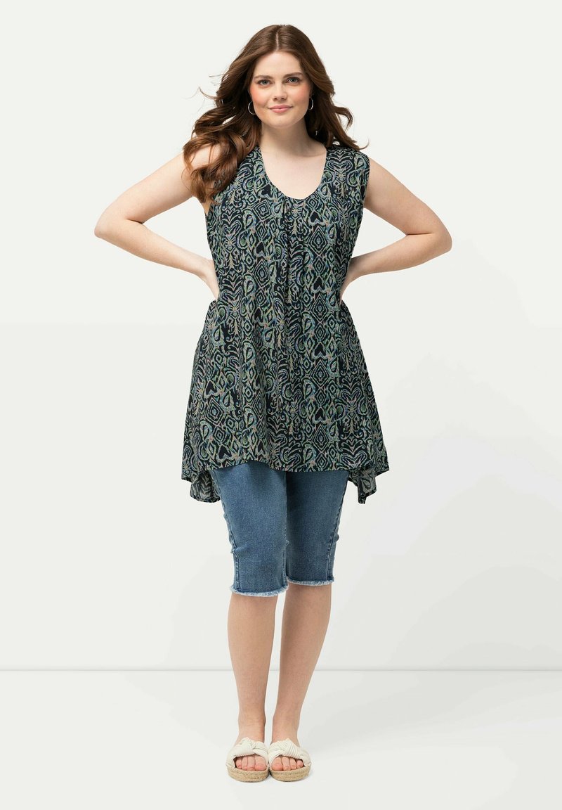 Mouwloze tuniek met een donkerblauw-groen bloemenpatroon, gecombineerd met knielange denim shorts. Lichtgekleurde sandalen maken de look af.