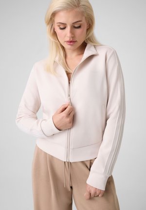 Femme blonde portant une veste zippée beige clair et un pantalon beige, tenant la fermeture éclair de la veste d'une main, regardant vers le bas.