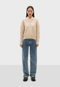 Pull en tricot crème avec un décolleté en V et des motifs géométriques texturés, associé à un jean bleu clair taille haute et des chaussures noires.
