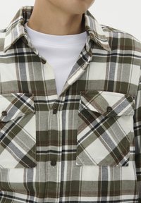 Zbliżenie na osobę w zielonej, brązowej i białej kratkowanej koszuli z guzikami noszonej na białej koszulce z okrągłym dekoltem.