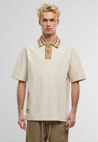 Beige Poloshirt mit einem strukturierten braunen Kragen mit weißen Streifen, zwei Knöpfen und einem Logopatch am Saum. Kurze Ärmel.