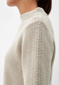 Beżowy sweter dzianinowy z wysokim kołnierzem, z fakturowanym ribbingiem i skośnym przeszyciem wzdłuż ramion. Miękka, fakturowana tkanina.