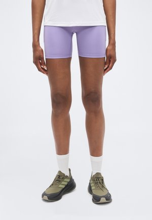 Person iført lilla sportsshorts, hvide sokker og olivengrønne Adidas-sneakers står mod en ensfarvet hvid baggrund.