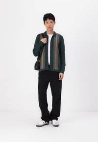 Cardigan verde scuro con motivo a strisce verticali beige, indossato sopra una camicia bianca, abbinato a pantaloni neri e sneakers bianche.