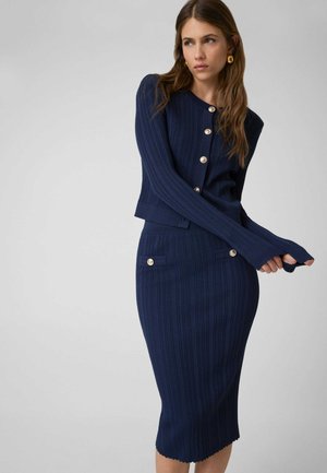 Femme portant un cardigan en maille côtelée bleu marine avec des boutons dorés et une jupe assortie, près du corps, avec des poches à boutons sur le devant.