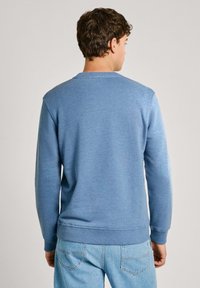 Mėlynas džemperis su ilgomis rankovėmis, apvaliu iškirpimu ir gipiūrinėmis apyrankėmis. Minkšta tekstūra ir minimalistinis dizainas, nešiojamas su šviesiai mėlynomis džinsinėmis kelnėmis.