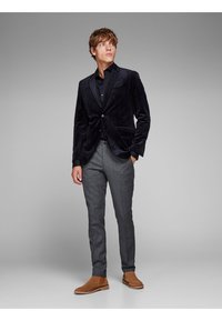 Marineblauwe fluwelen blazer met pieklabel, grijs geruite broek en bruine chelsea boots. Zwarte shirt onder de blazer; zakken zichtbaar.