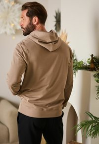 Beige hættetrøje med snørelukning i hætten, blød tekstur, tætsiddende design og ribbede manchetter og kant. Bæret med sorte bukser foran en neutral baggrund.