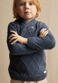 Blauwe gebreide trui met ruitpatroon, geribde kraag en mouwen, voorzien van houten knopen. Model met gekruiste armen, broeken niet zichtbaar.