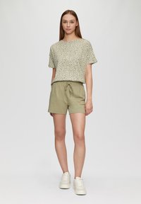 Korte mouwen, gespikkeld groene top gecombineerd met olijfgroene shorts met een elastische tailleband en trekkoord, gedragen met witte sneakers.