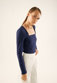 Granatowa bluzka z długim rękawem, kwadratowym dekoltem i dopasowanym gorsetem, zestawiona z białymi, wysokimi spodniami. Gładka faktura, minimalistyczny design.