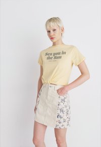Gul t-skjorte med teksten "Sea you in the Sun", kombinert med en kremfarget denimskjørt med blomsterdesign på den ene siden.