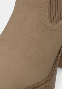 Boots de gleznă din suede maro, cu panouri laterale elastice texturate și un toc ușor curbat. Finisaj neted cu un luciu subtil.