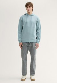 Felpa di colore azzurro con tasca a canguro e cordino. Indossata con jeans grigi e sneakers bianche. Presenta un marchio discreto sul petto.