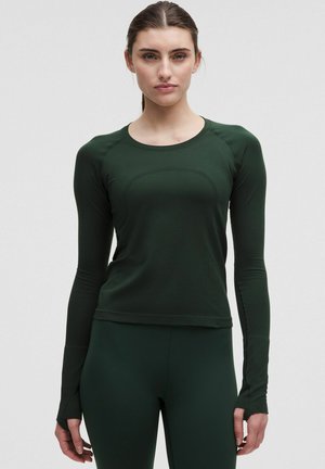 Noor naine kannab tumerohelise pikakäistega sporditoppi, millel on sõrmeaugu ja sellele sobivaid leggins’e, seistes ühetaolise heleda tausta ees.