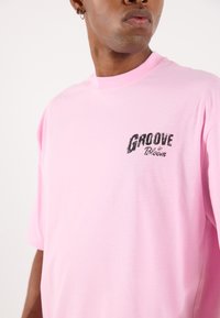 Pink cotton T-shirt with a relaxed fit. Features "GROOVE by Bloom" printed in bold black letters on the left chest. Short sleeves, smooth texture.
