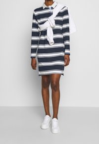 Gestreiftes Hemdkleid in Marineblau und Weiß mit langen Ärmeln und Kragen, kombiniert mit weißen Sneakers. Ein Pullover um die Schultern gebunden als Akzent.