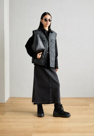 Black quilted vest over a black shirt, paired with a long denim skirt and black chunky boots. Accessory includes a black bag and sunglasses.