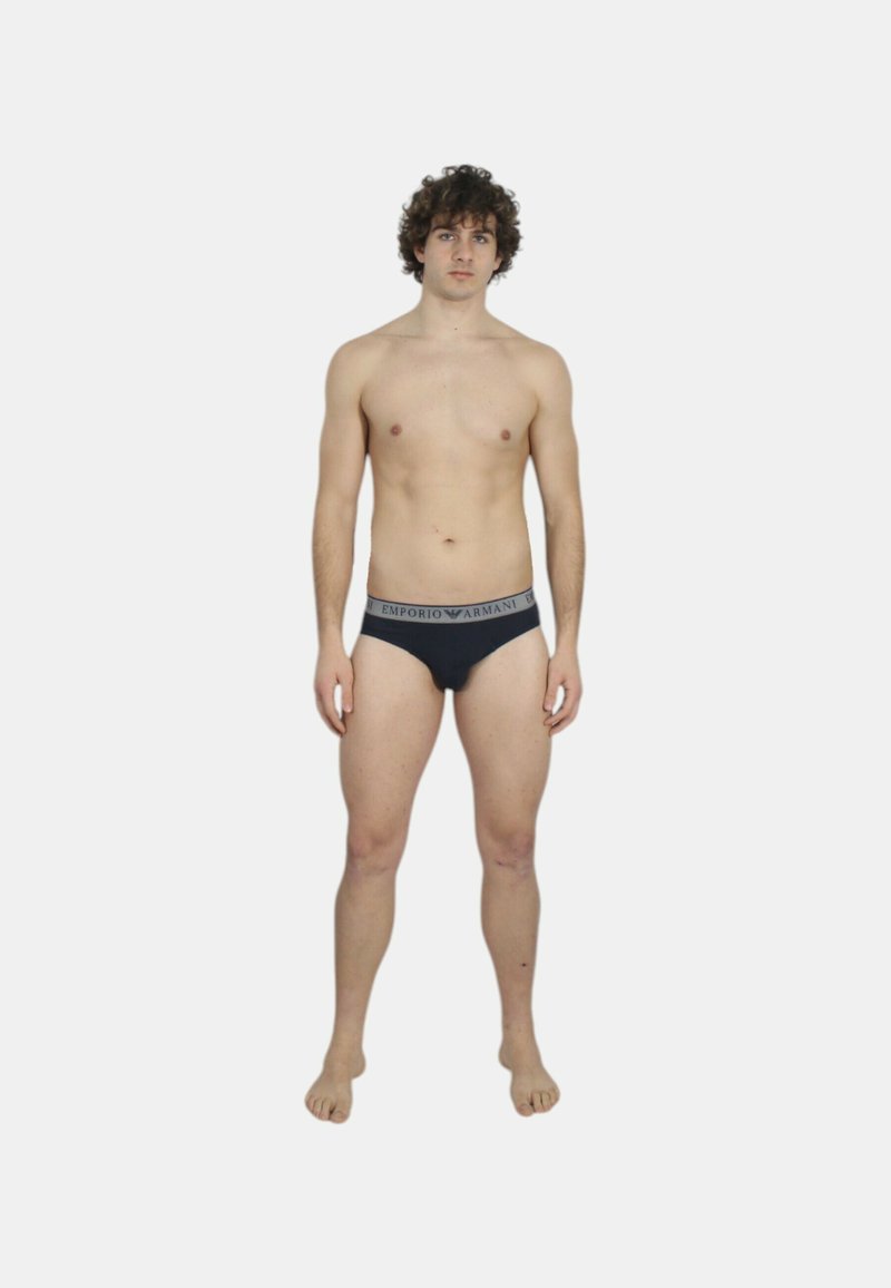 Slip nero con fascia grigia in vita con logo "Emporio Armani". Il modello è in piedi, a piedi nudi, mostrando un design e una vestibilità minimal.