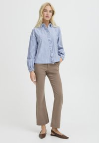 Moteris vilkinti mėlynai ir baltai dryžuotą raukinį palaidinę, smėlio spalvos tiesias kelnes ir rudas smailias slingback basutes, stovi prieš baltą foną.