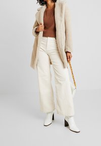 Beige Kunstfellmantel mit Kapuze, brauner Strickpullover, cremefarbene Cord-Hose mit weitem Bein und weiße Stiefeletten mit Absatz.