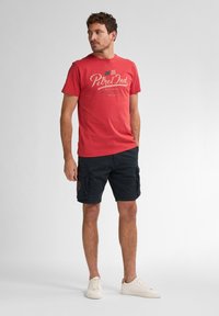 Rød bomulds t-shirt med "Petrol Ind" grafik, parret med marineblå cargo shorts; modellen har hvide sneakers på og står mod en lys baggrund.