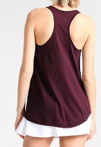 Débardeur bordeaux à dos nageur, en tissu doux avec un ourlet courbé. Porté sur une jupe blanche, mettant en valeur un look superposé.