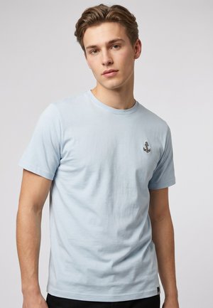 Junger Mann mit hellbraunen Haaren, der ein hellblaues T-Shirt mit kleinem Anker-Emblem auf der linken Brust vor einem einfarbigen Hintergrund trägt.