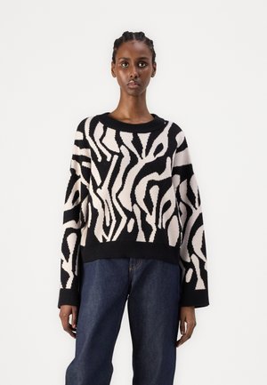 Pull en tricot noir et crème avec un motif abstrait, col rond et manches longues. Porté sur un pantalon en denim bleu, coupe décontractée.