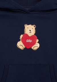 Φούτερ με κουκούλα σε ναυτικό μπλε χρώμα με εικόνα αρκούδας που κρατάει μια κόκκινη καρδιά. Η καρδιά έχει εκτυπωμένο το "GAP" με λευκά γράμματα. Μαλακό ύφασμα με τσέπη μπροστά.