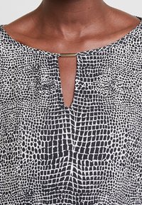 Czarnobiały wzorzysty top z teksturowanym wzorem, dekoltem w serek z małym otworem i złotym akcentem przy rozcięciu.