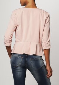 Vaaleanpunainen peplum-takki, jossa on kolmivartiset hihat, sileä kangas ja rypytysdetailit hihoissa, yhdistetty tummansinisiin farkkuihin.