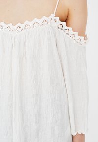 White off-the-shoulder top featuring a textured fabric, scalloped lace trim along the neckline and sleeves, and loose-fitting design.